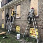 People painting boarded windows on building exterior.