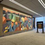 A colorful mural featuring abstract human and animal figures on an urban underpass wall, with modern lighting overhead.