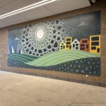 A colorful mural depicting an abstract landscape with sun, hills, houses, and a patterned sky, installed on an urban building's exterior wall.