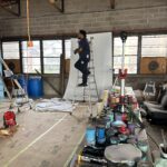 A person on a ladder working in a cluttered workshop with painting supplies and equipment.