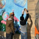 Two individuals installing a panel of artwork on an urban building exterior.